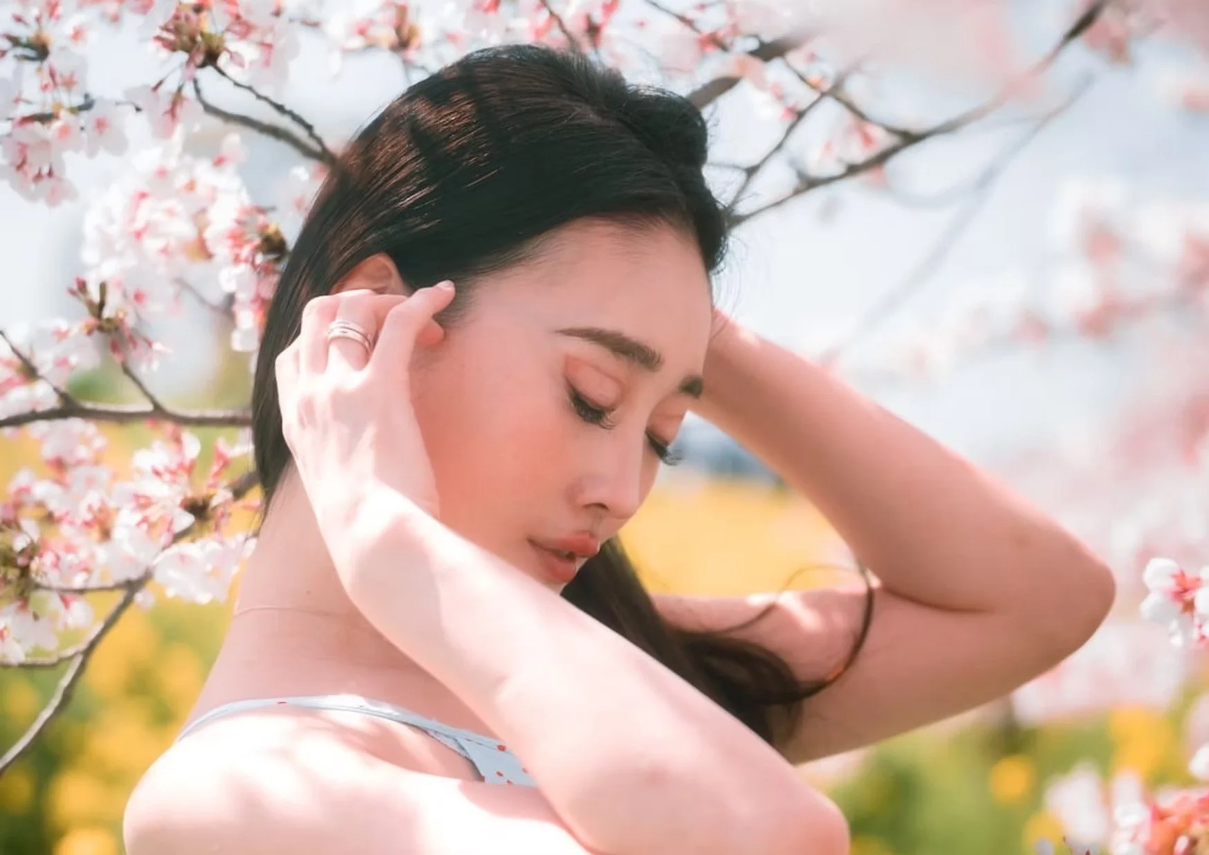 A woman in a light blue dress stands among blooming cherry blossoms, gently touching her hair. The soft sunlight and vibrant pink flowers create a serene, springtime atmosphere.