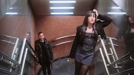 A woman with long dark hair wearing a black leather jacket and skirt stands at the top of a staircase with two men at the bottom, in a modern indoor space with brown walls and metallic railings.
