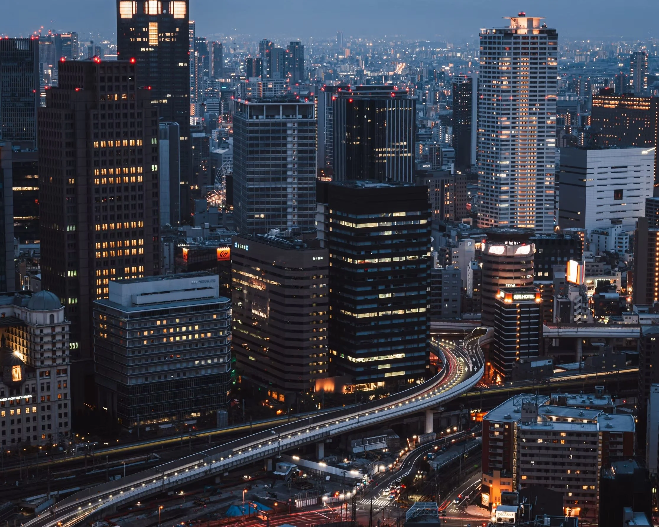 A city skyline at dusk with illuminated skyscrapers, office buildings, and highways with streaks of car lights.