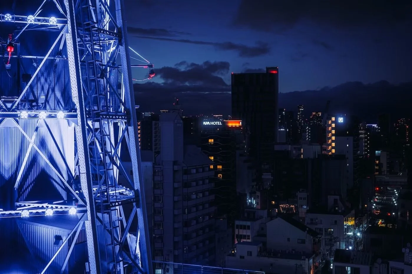 Nighttime cityscape with illuminated high-rise buildings and a Ferris wheel structure with bright blue lights in the foreground. Tsutenkaku tower in Shinsekai Osaka 