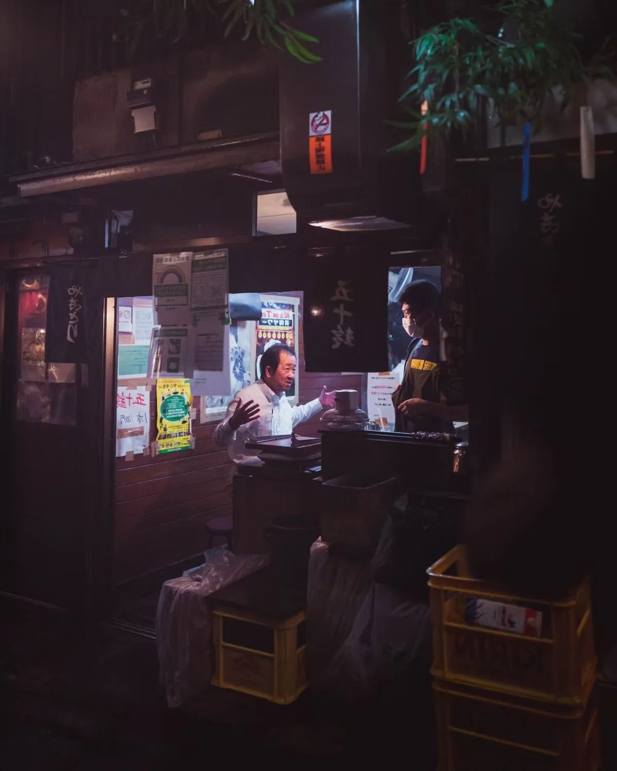 A man in a white shirt engaged in conversation with a person wearing a face mask inside a small, dimly-lit restaurant or food stall, with various signs and papers on the wall behind them. Shot in Omoide Yokocho.