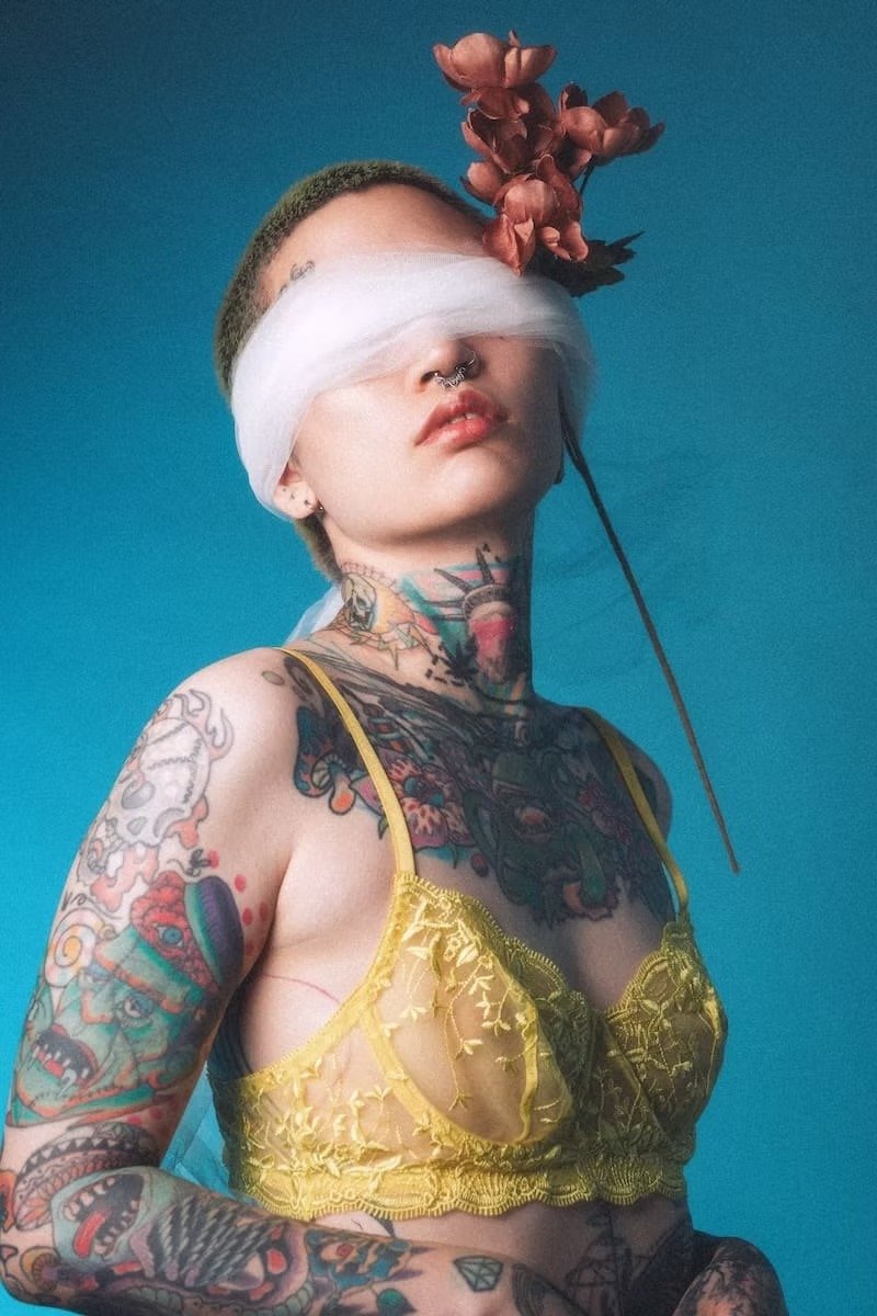 A tattooed woman with colorful tattoos, wearing a yellow lace top, blindfolded with white fabric, with a bunch of pink flowers on her head against a blue background. Shot in Tekumi Studios.