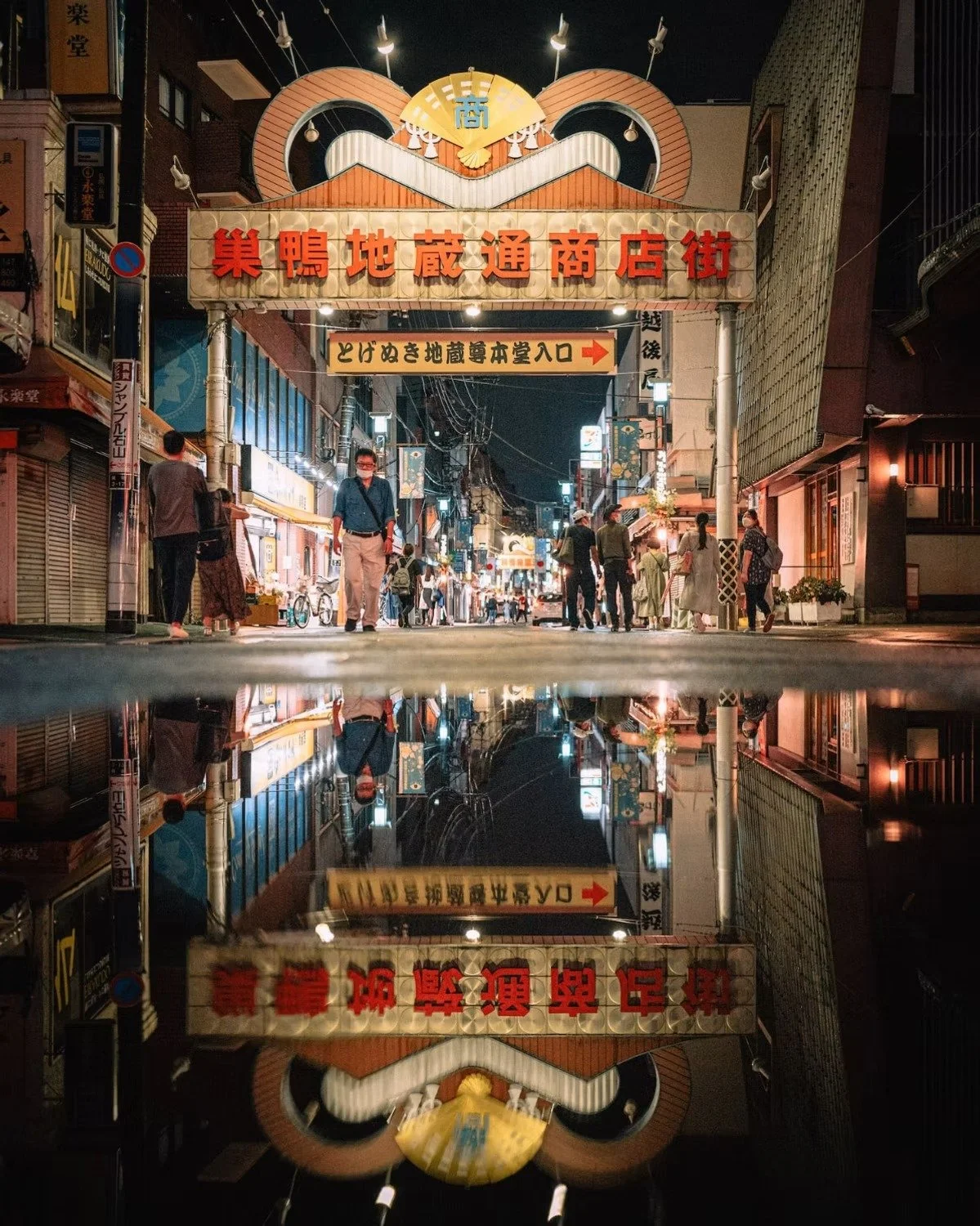 A lively night scene of a Japanese shopping street with illuminated signs and people walking. The street is reflected in a puddle on the ground, creating a mirror image.