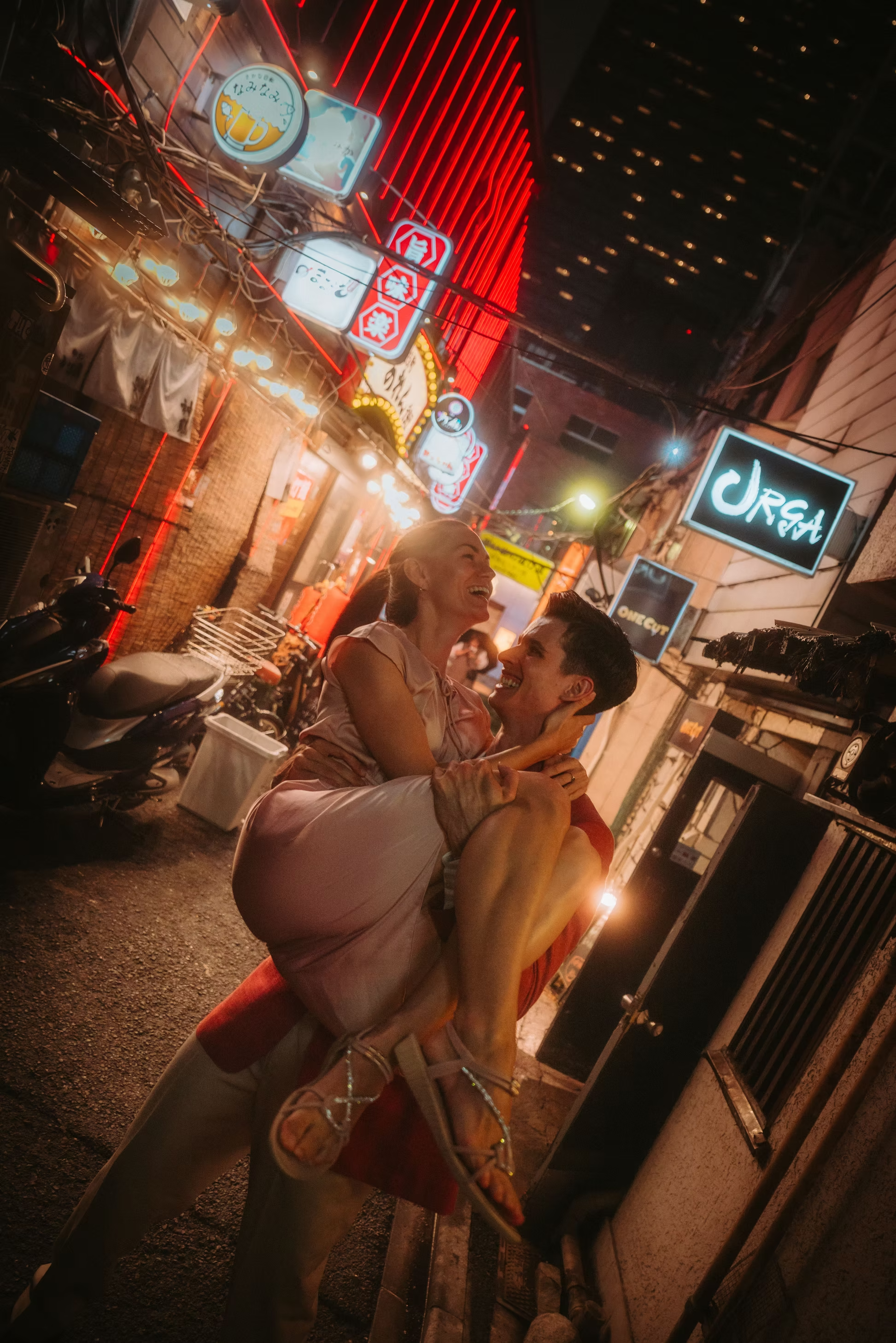 A couple enjoying a night out on a lively street with neon signs, one showing beer, another with Japanese characters, and various other signs. The woman is sitting on the man's lap, both smiling and laughing amid warm street lighting and a vibrant city atmosphere.