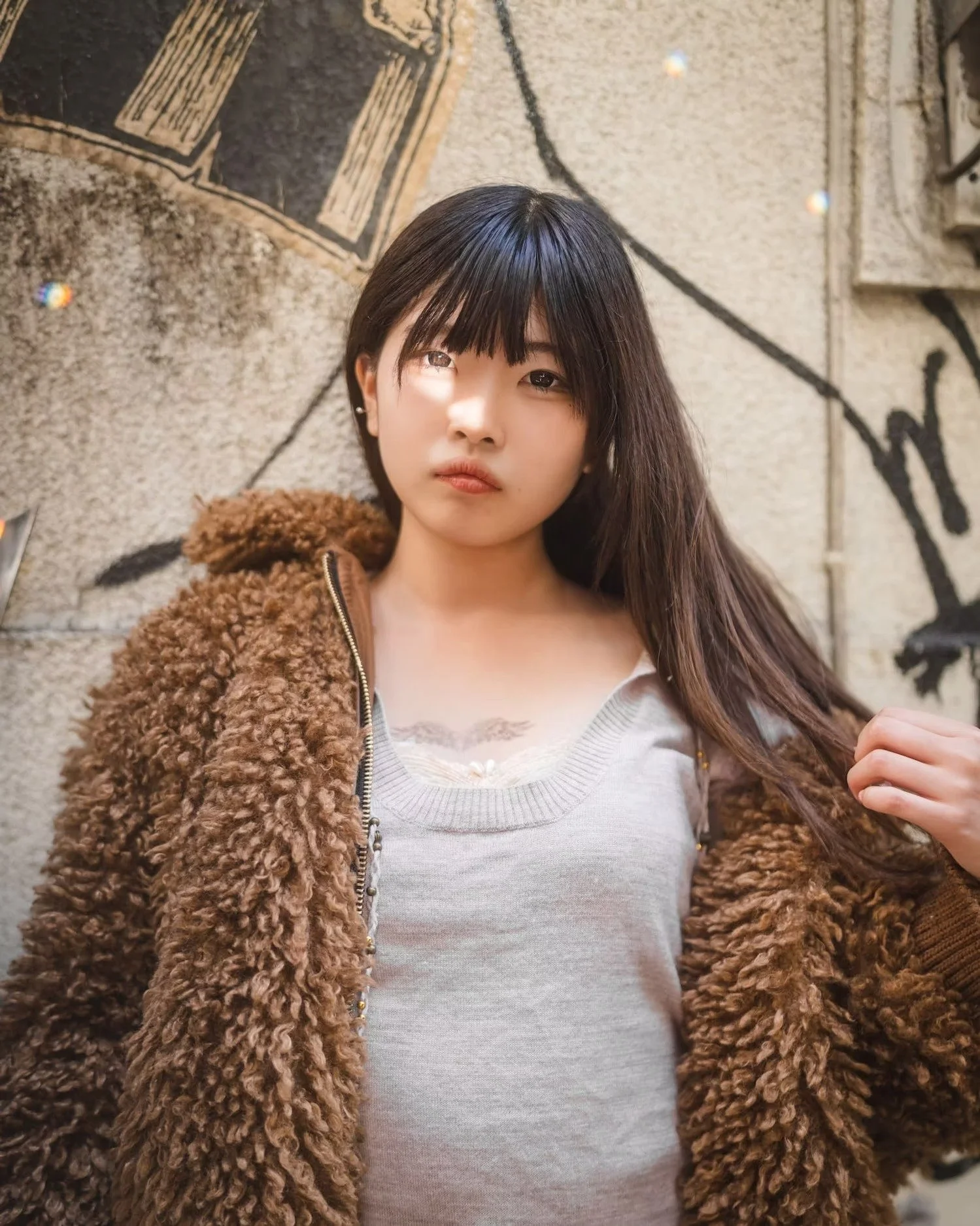 A young woman with straight dark hair and bangs, wearing a white top and a brown furry jacket, poses against a textured concrete wall with black graffiti.