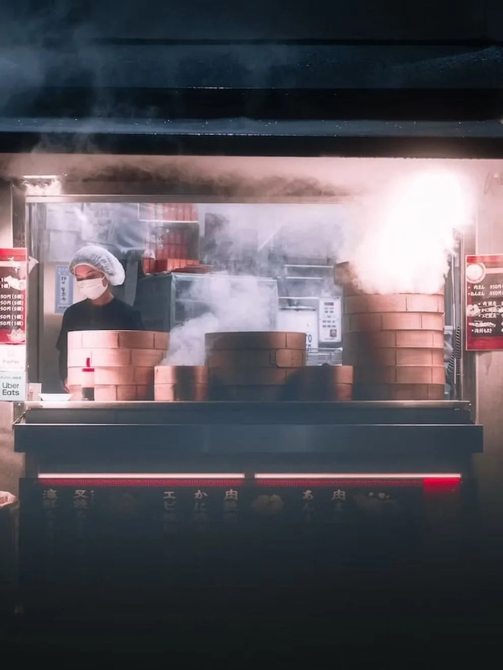 A street food vendor preparing food behind a glass display, with steam rising from stacked steaming baskets. The vendor wears a hairnet and face mask, with a red straw protruding from a drink in front. The scene appears to be in a busy commercial kitchen or food stall.