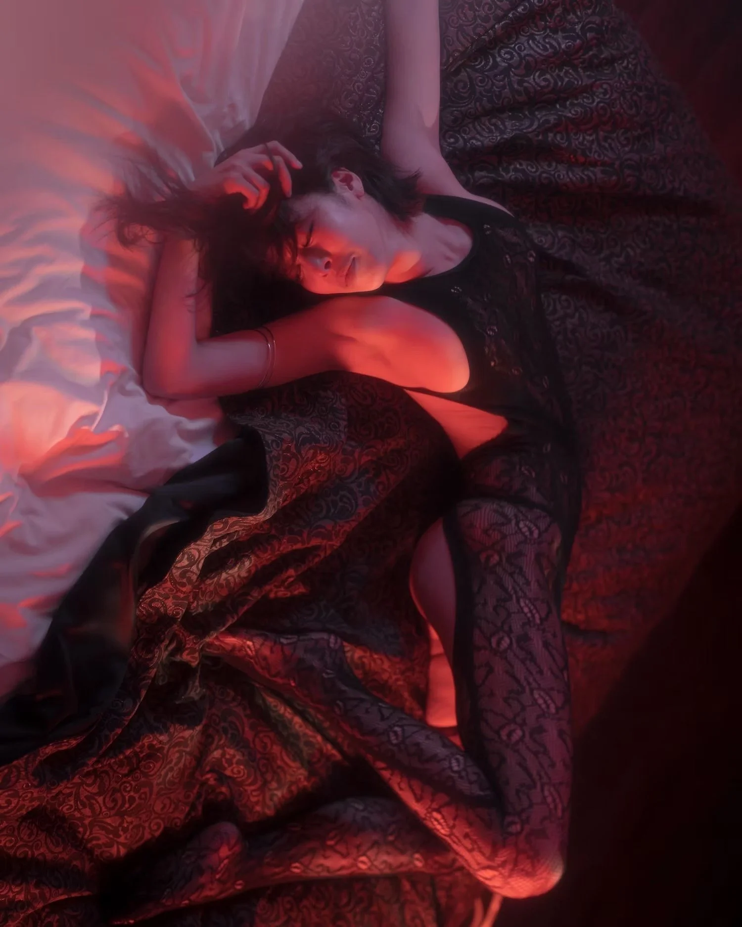 A woman with dark hair and closed eyes lies on a bed with patterned blankets and white pillows, illuminated by red lighting.