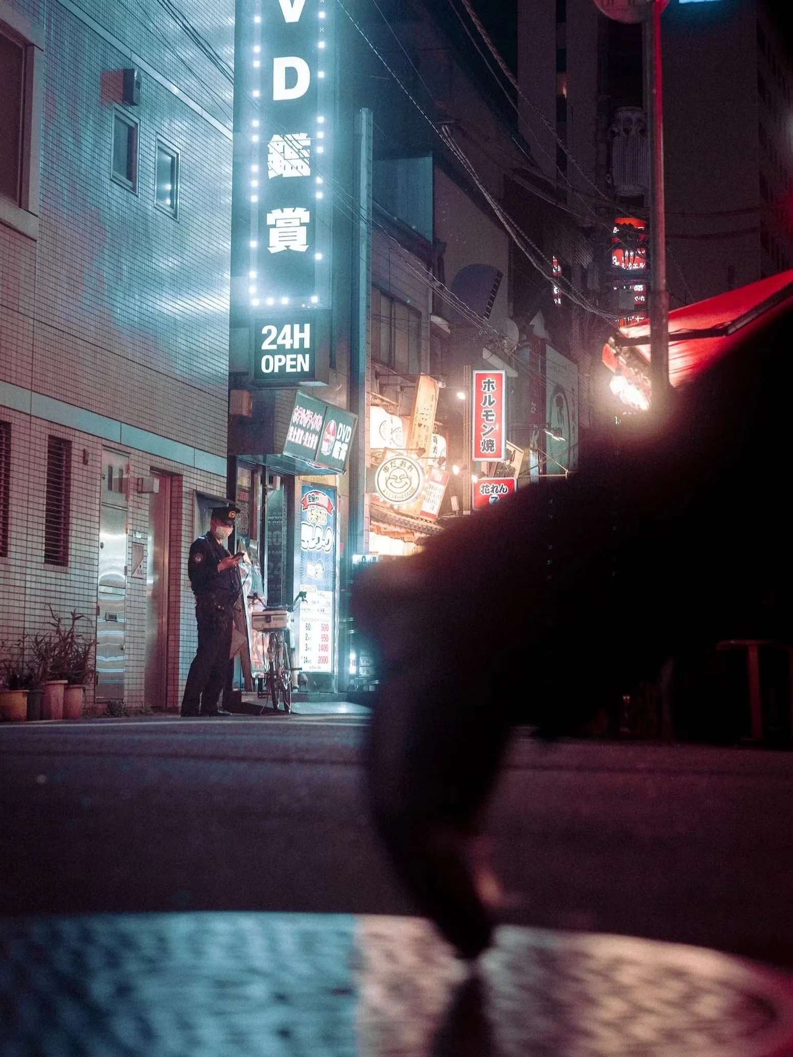 Nighttime street scene with illuminated signs in Japanese, a man wearing a mask standing next to a bicycle, and a row of buildings with various advertisements and lights.
