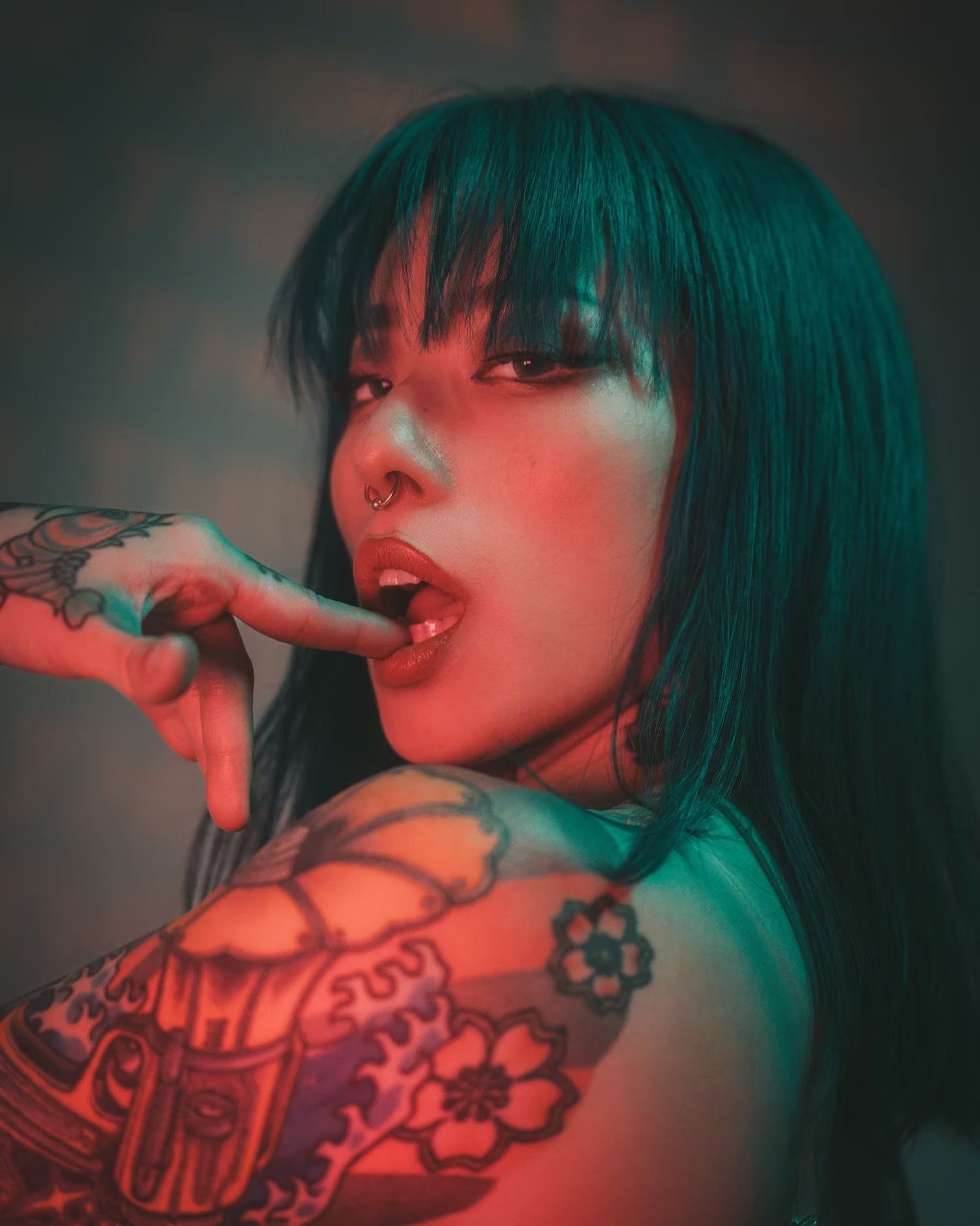 A woman with teal-colored hair, tattoos on her arm, and a septum piercing poses with her finger near her mouth, in a dimly lit environment with red and green lighting.