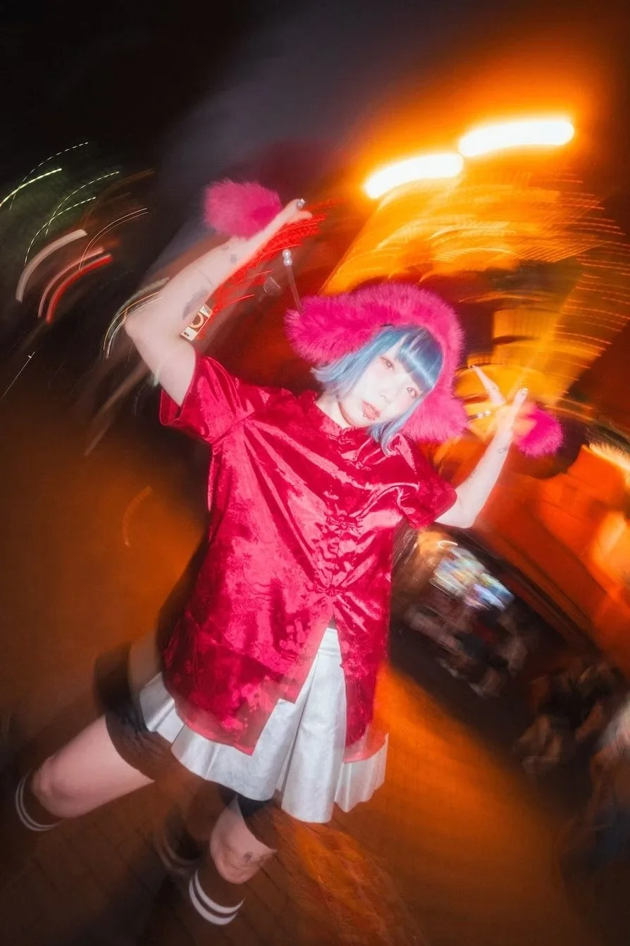 A young Japanese woman with blue hair and bangs, wearing a pink furry hood, a red velvet top, a beige skirt, and striped socks, posing with her arms raised in a nighttime city street with streaks of orange and yellow lights in the background. Shot by