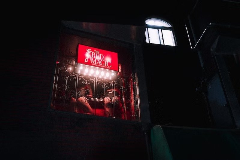 A neon-lit sign reading 'The Red Magic' above a window with red curtains and decorative metalwork, in a dark interior setting. Shot on the Tokyo Redlights street photography workshop by Tokyo street photographer Deniz Demir.