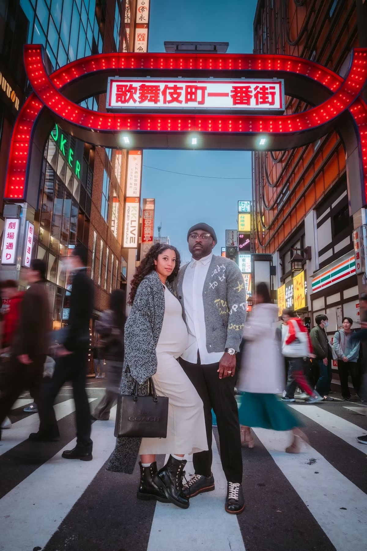 A couple stands on a busy city street crosswalk in front of illuminated signs and buildings, with a colorful urban background during evening time.