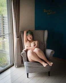 A woman sitting topless in a checkered armchair next to a window with curtains, with her arms and legs crossed.