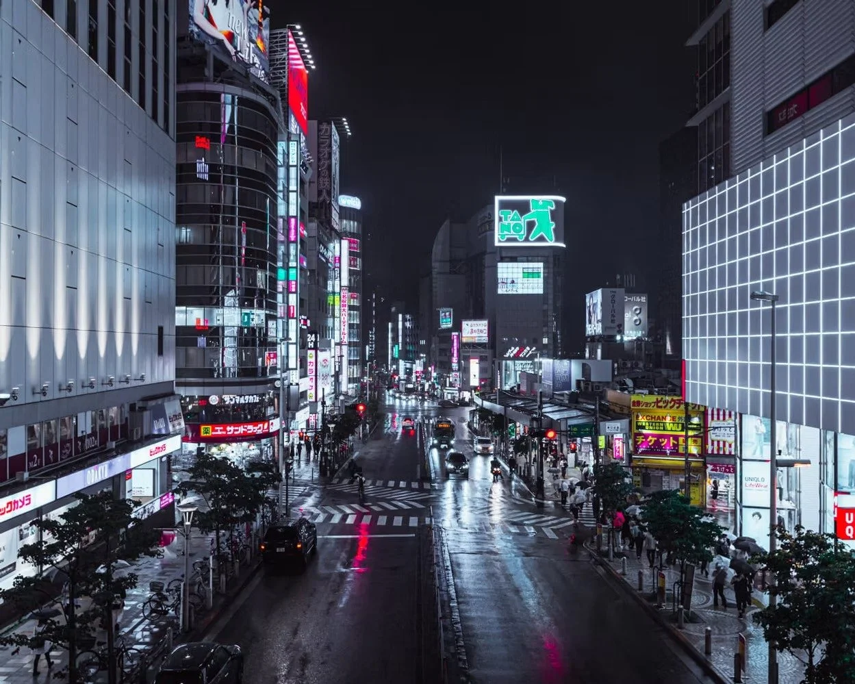 Nighttime city street scene with bright neon signs, wet pavement, and people with umbrellas.