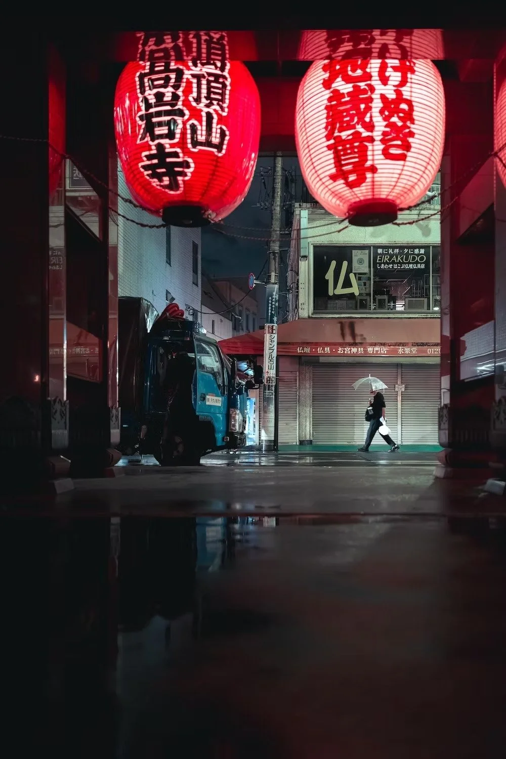 Night scene of a Japanese street featuring red paper lanterns hanging above, a sidewalk with a person holding an umbrella walking past closed shops, and a small blue truck parked on the side. Shot by Tokyo street photographer Deniz Demir.