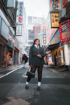 A woman in a black suit and skirt stands confidently on a Tokyo street, surrounded by colorful neon signs and tall buildings. She carries a black bag and wears knee-high socks and shoes.