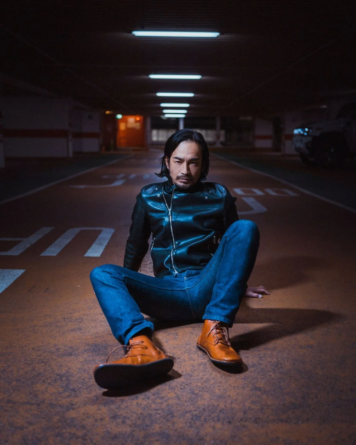 A man with long dark hair and facial hair sitting on the ground in an underground parking garage at night, wearing a black leather jacket, blue jeans, and orange boots, with a serious expression.