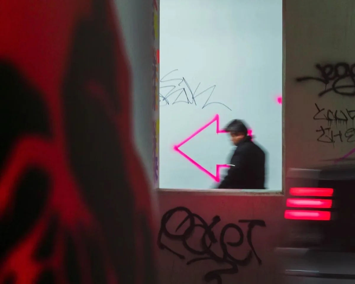 A person wearing a hat and black coat walking past a window with graffiti on the walls, pink arrow neon lights on the wall in the background shot in Shibuya by Tokyo street photographer Deniz Demir.