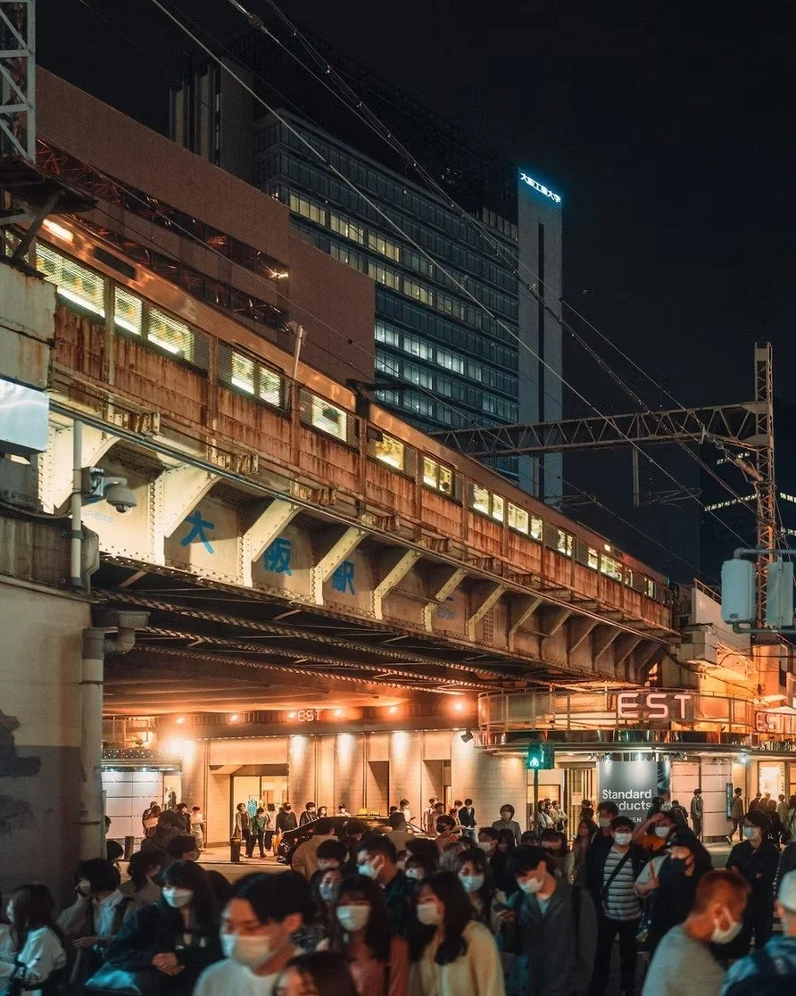 Photographed by Tokyo photographer Deniz Demir a crowd of people wearing masks on a city street at night under an elevated train track with high-rise buildings in the background.