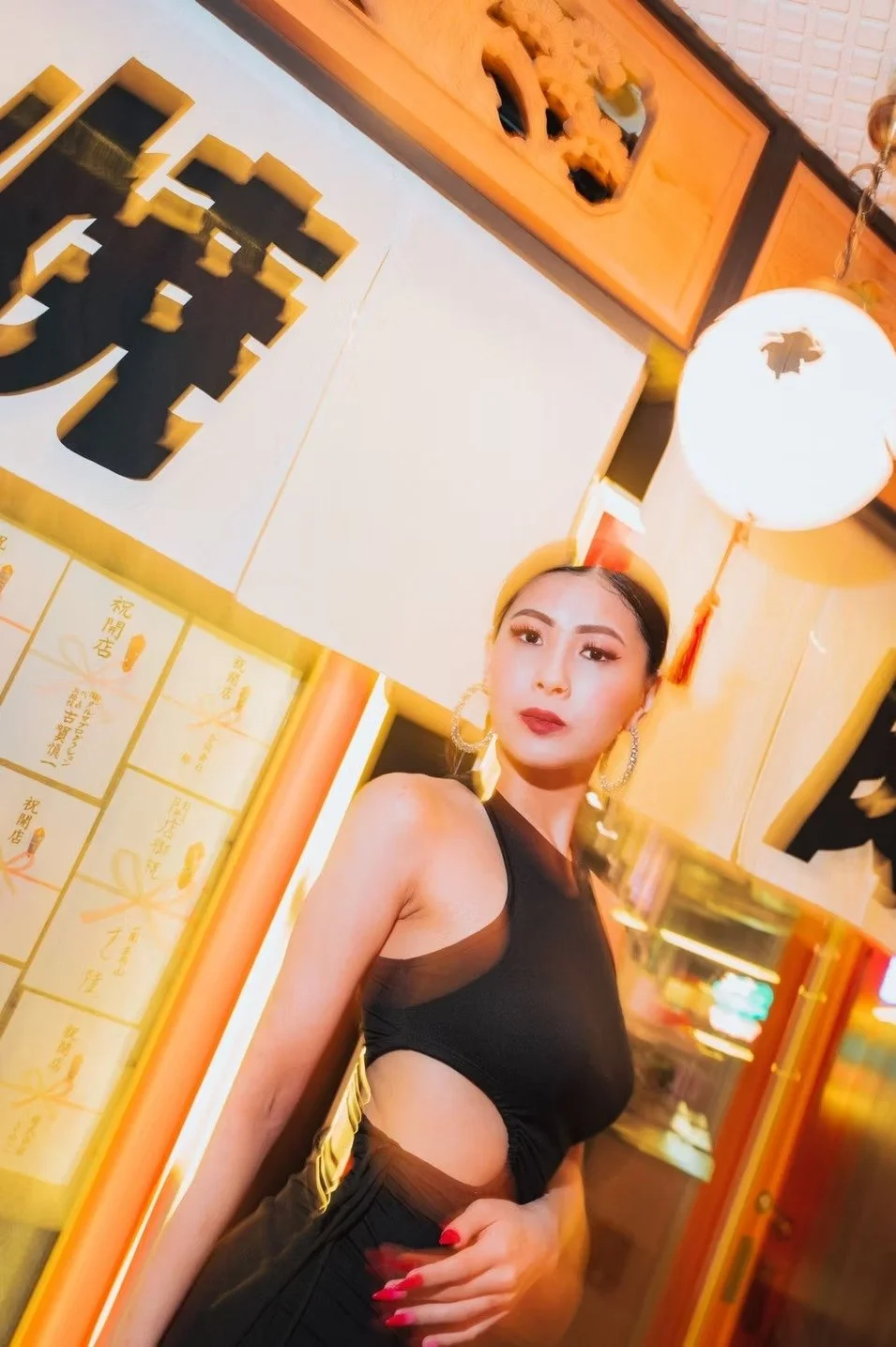 A woman in a sleek black cut-out dress stands confidently in front of a traditional Japanese storefront adorned with lanterns and gold accents. The vibrant lighting enhances the modern-meets-traditional vibe. Street photography by Deniz Demir.