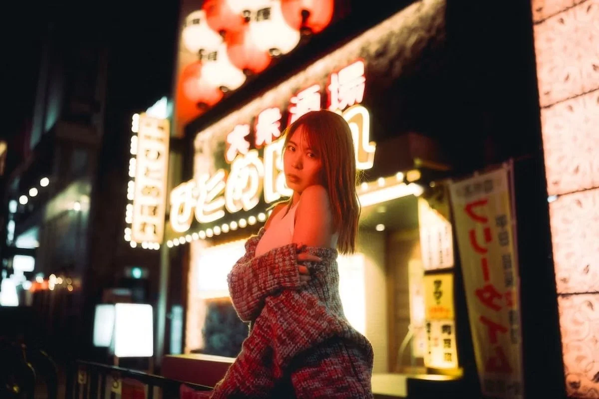Portrait photography of a young woman posing at night in front of a brightly lit sign with Asian characters and lanterns, wearing a coat draped off her shoulders.