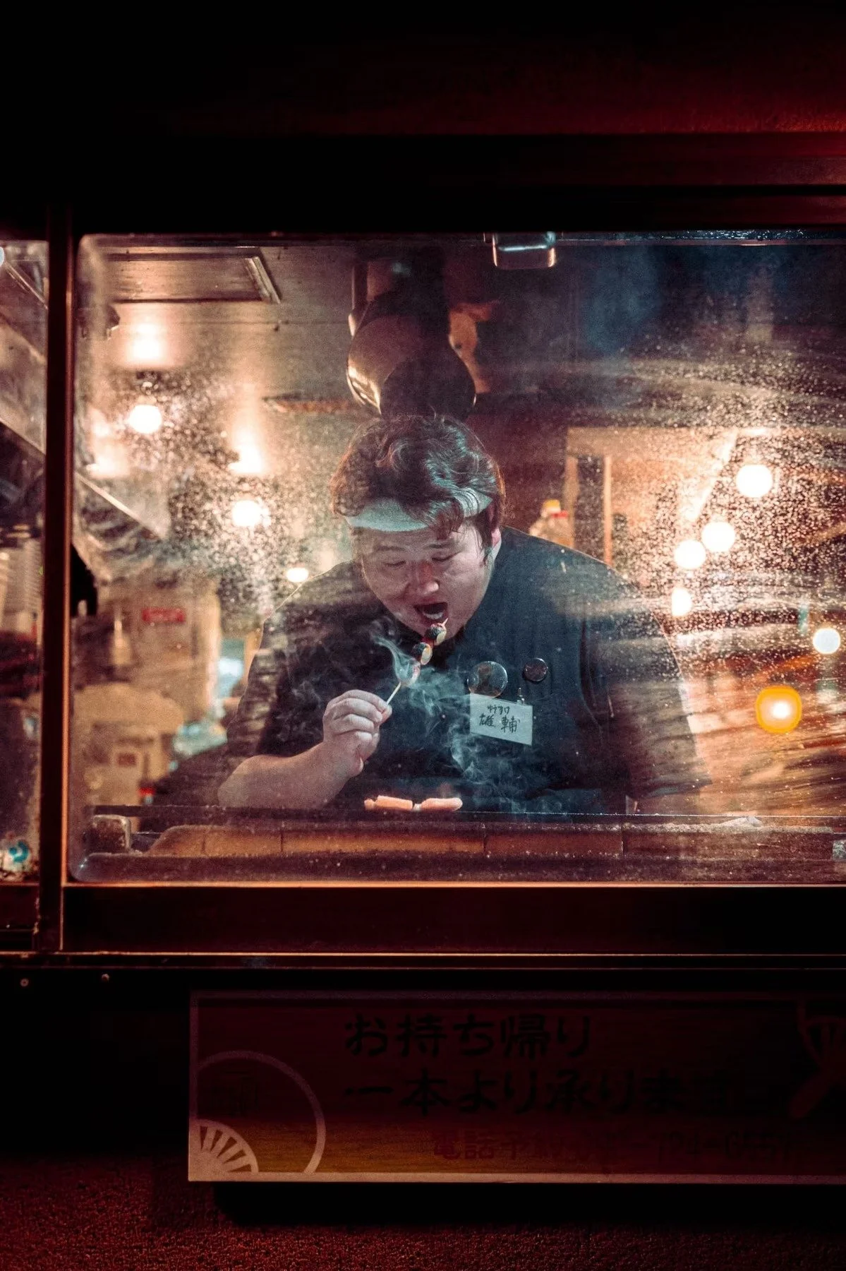 A Japanese person with a headband is making a bubble with a bubble wand behind a glass window, with reflections of lights and some objects around in the background.