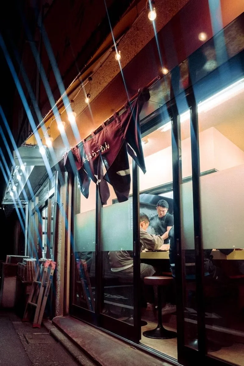 View of a Japanese ramen restaurant with customers inside, visible through glass windows, and traditional fabric wall hanging.