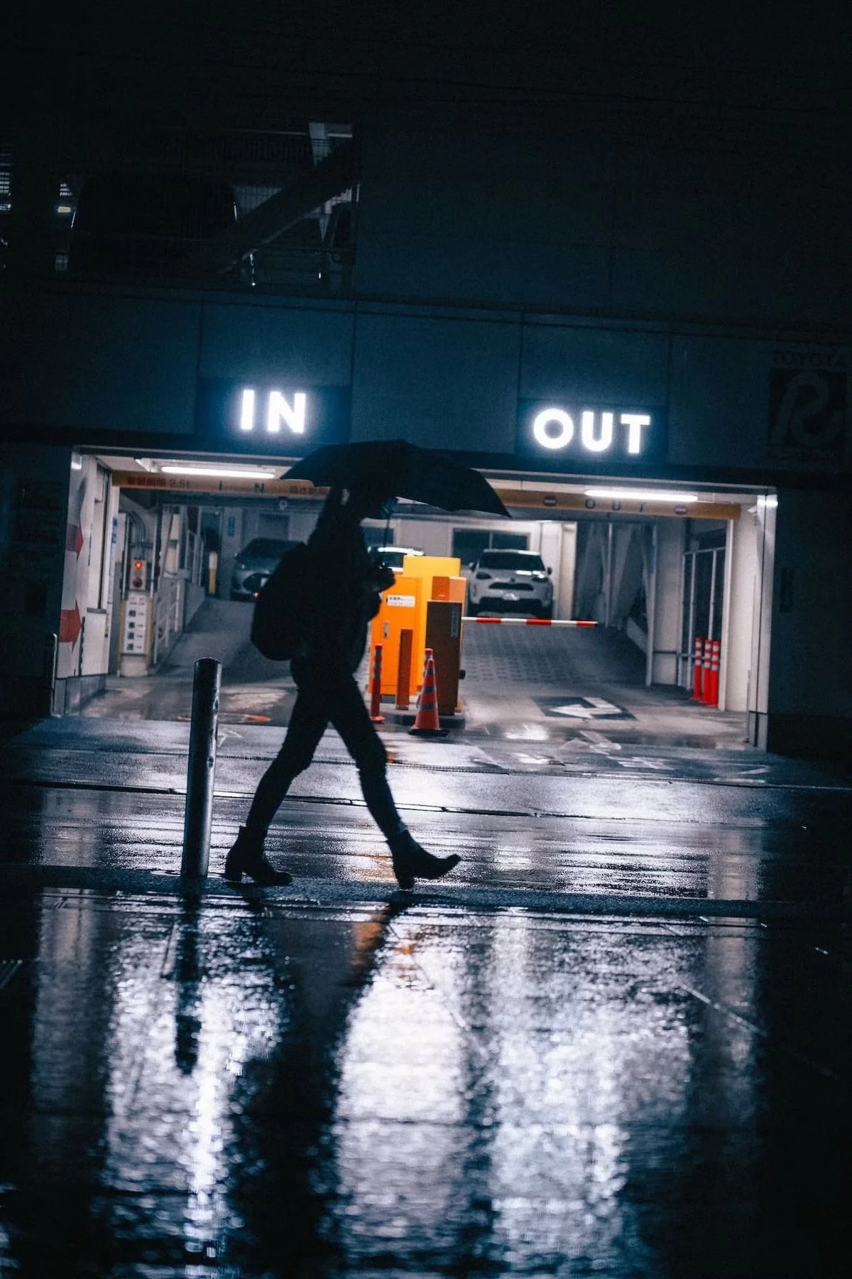 A person walking with an umbrella near an entry and exit parking garage in rainy weather at night, with wet reflective ground shot in Shibuya Tokyo.