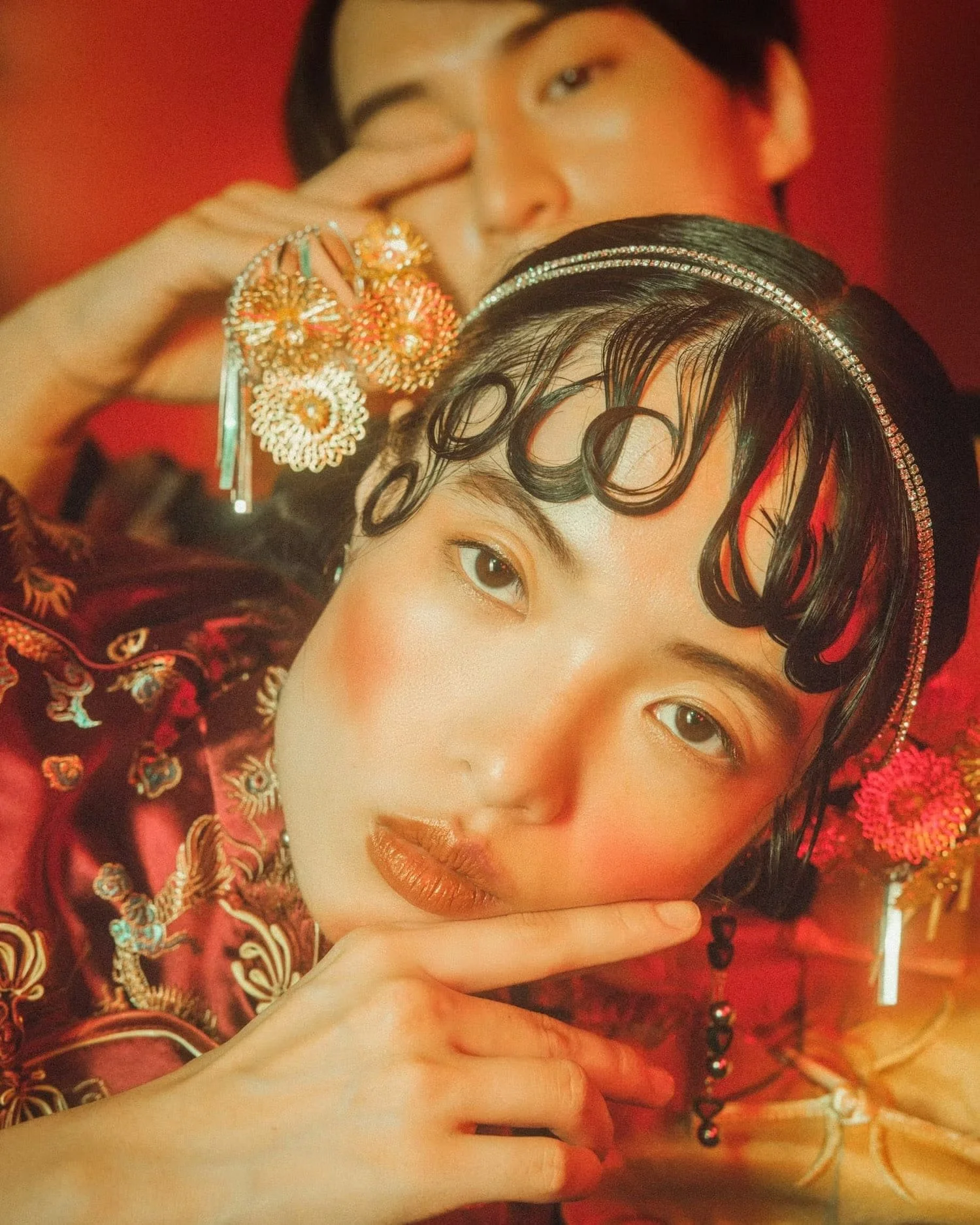 Close-up of a woman with ornate jewelry and makeup, resting her chin on her hand, wearing a red embroidered garment, with a blurred red background.