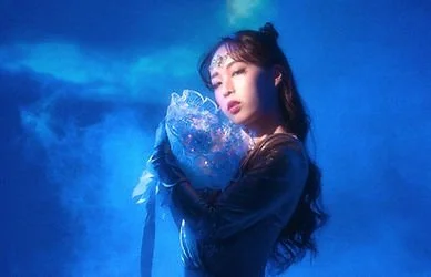 A woman in dark clothing holding a large, glittery, heart-shaped object against a blue background.