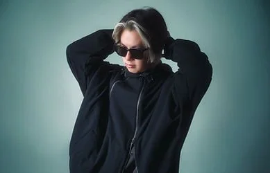 Person with short blonde hair wearing sunglasses and a black jacket, posing with hands behind head against a gradient green background.