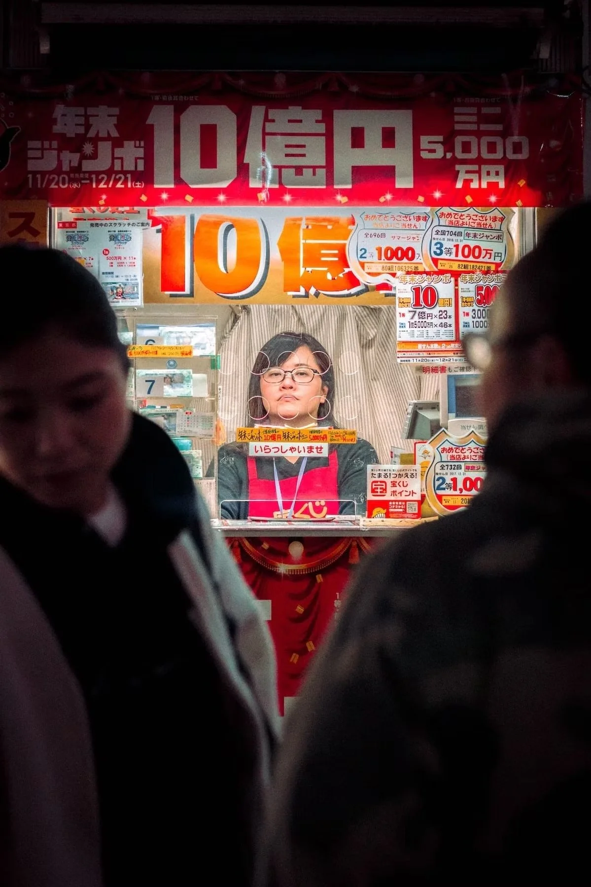 View through a glass window of a lottery ticket booth with a woman standing behind the counter, surrounded by various signs and posters. Two people are partially visible in the foreground on either side. Shot in Shibuya.