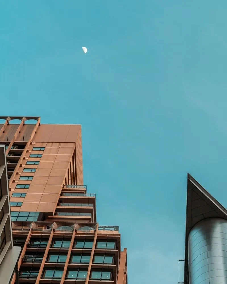 View of tall modern buildings against a clear blue sky with a visible moon in the upper part of the image.