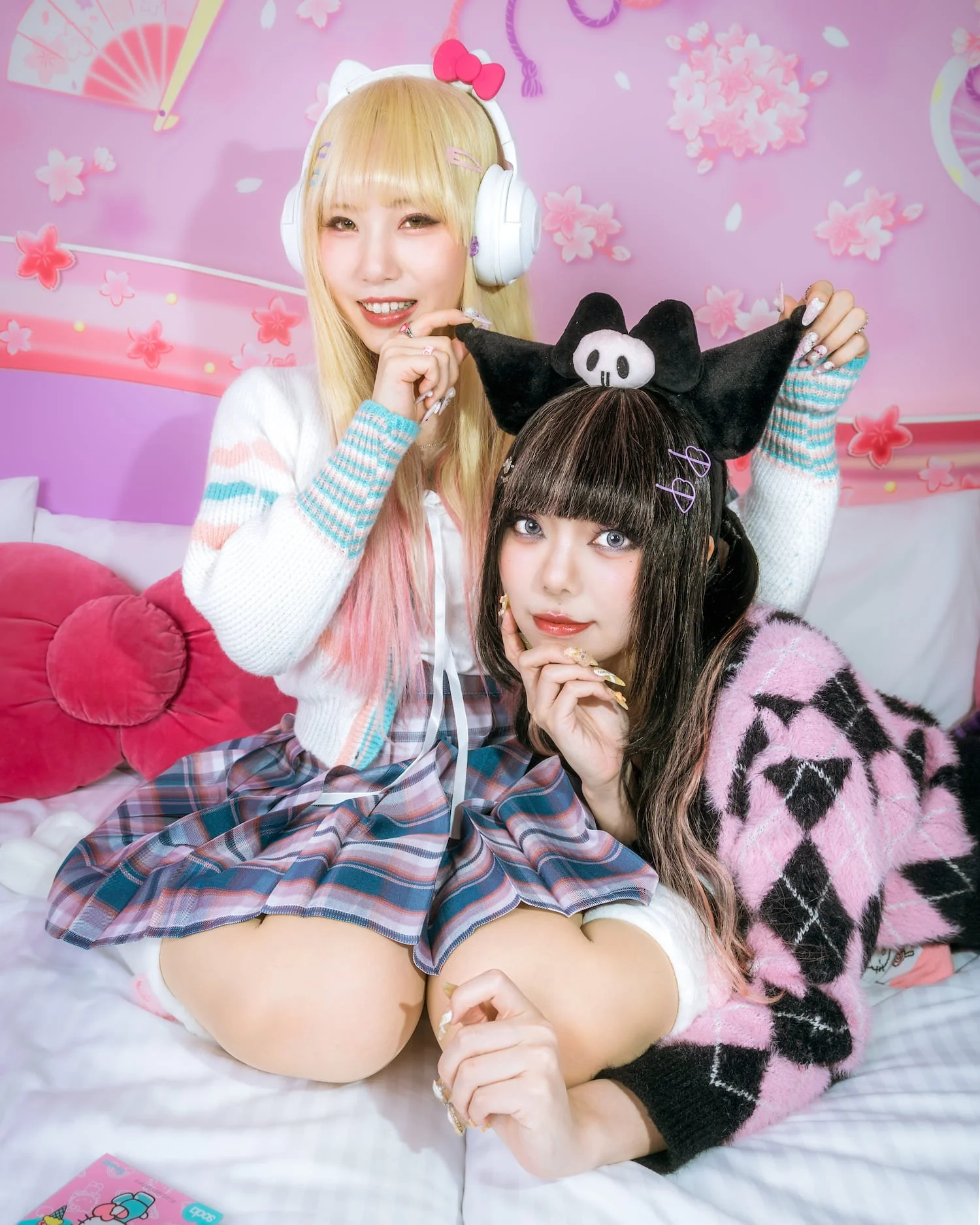 Two young women with colorful and playful outfits, one wearing a pink and black fleece with a pattern, and the other with blonde hair, cat ear headphones, and a pastel sweater, posing in a pink-themed room with cherry blossoms and a bed with pink pillows.