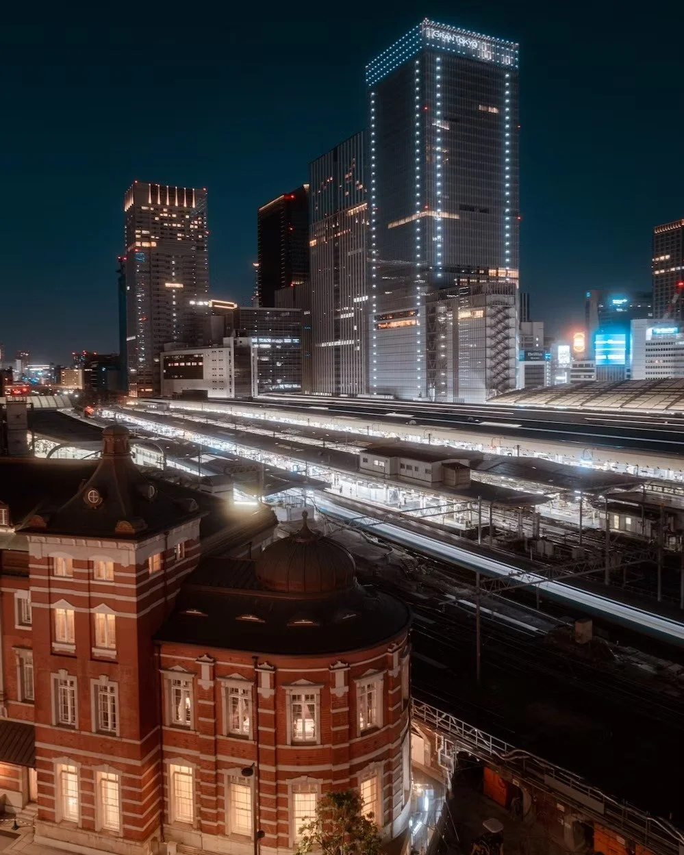 Nighttime cityscape featuring modern high-rise buildings with bright lights, illuminated train tracks, and a historic brick building in the foreground.