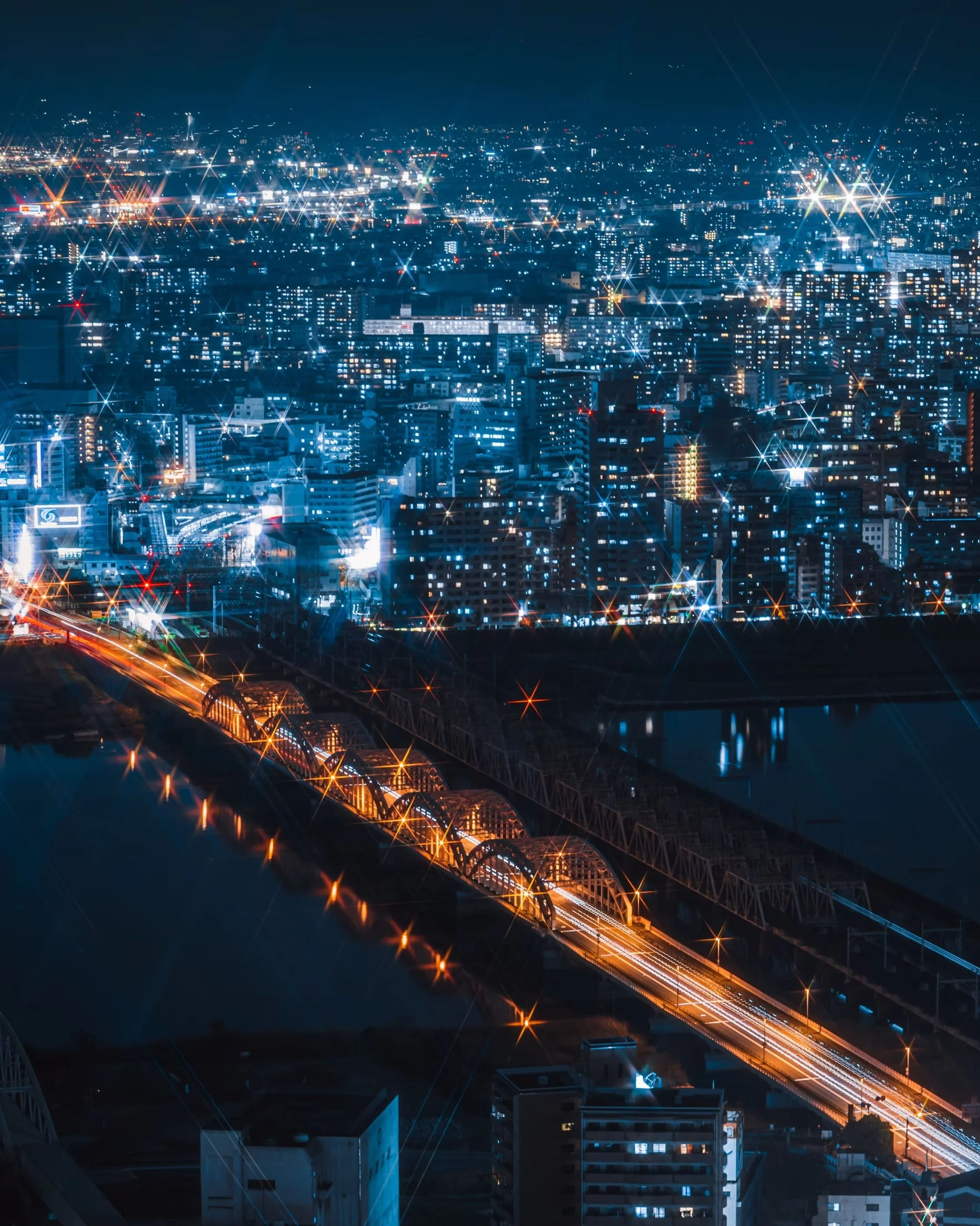 Night cityscape with illuminated buildings and roads, bright star-like lights on the buildings and streetlights, reflecting on water below.