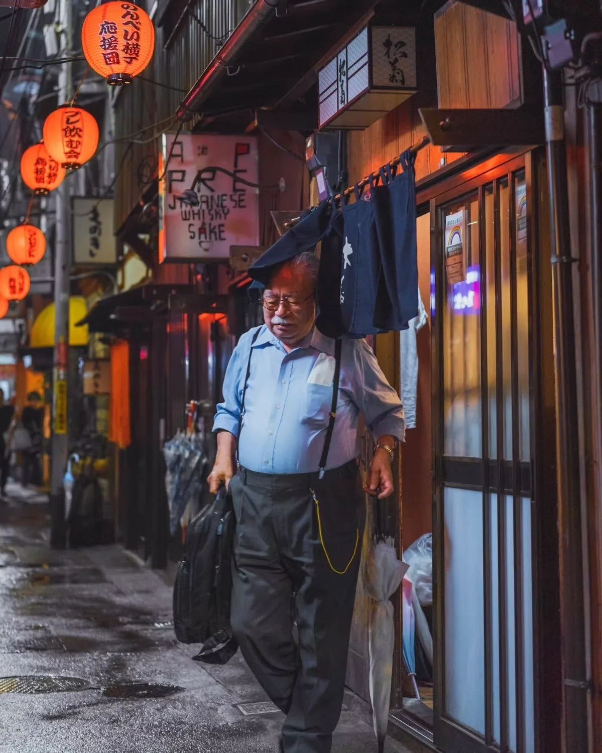 A Japanese man in a light blue shirt and dark pants walking on a narrow alleyway at night, carrying a black bag in his right hand, with neon and paper lantern lights illuminating the street, and signs in Japanese characters overhead.