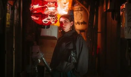 A young woman with makeup and earrings standing in a dimly lit corridor with red and orange lights and paper lanterns hanging from the ceiling.
