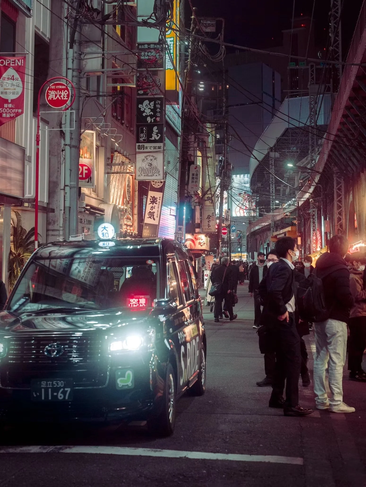 Night scene of a busy city street with numerous illuminated signs in Japanese, people walking, and a black van in the foreground.