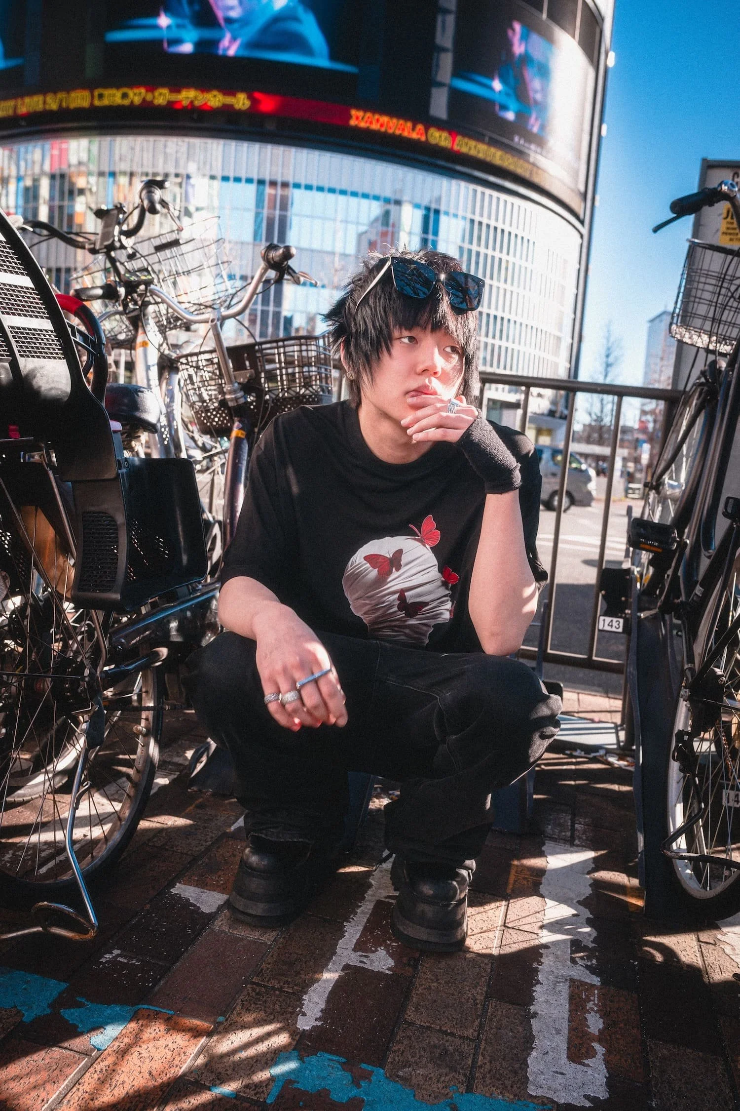 Person squatting in front of bicycles on a city street, with a large electronic billboard displaying a person and text in the background.
