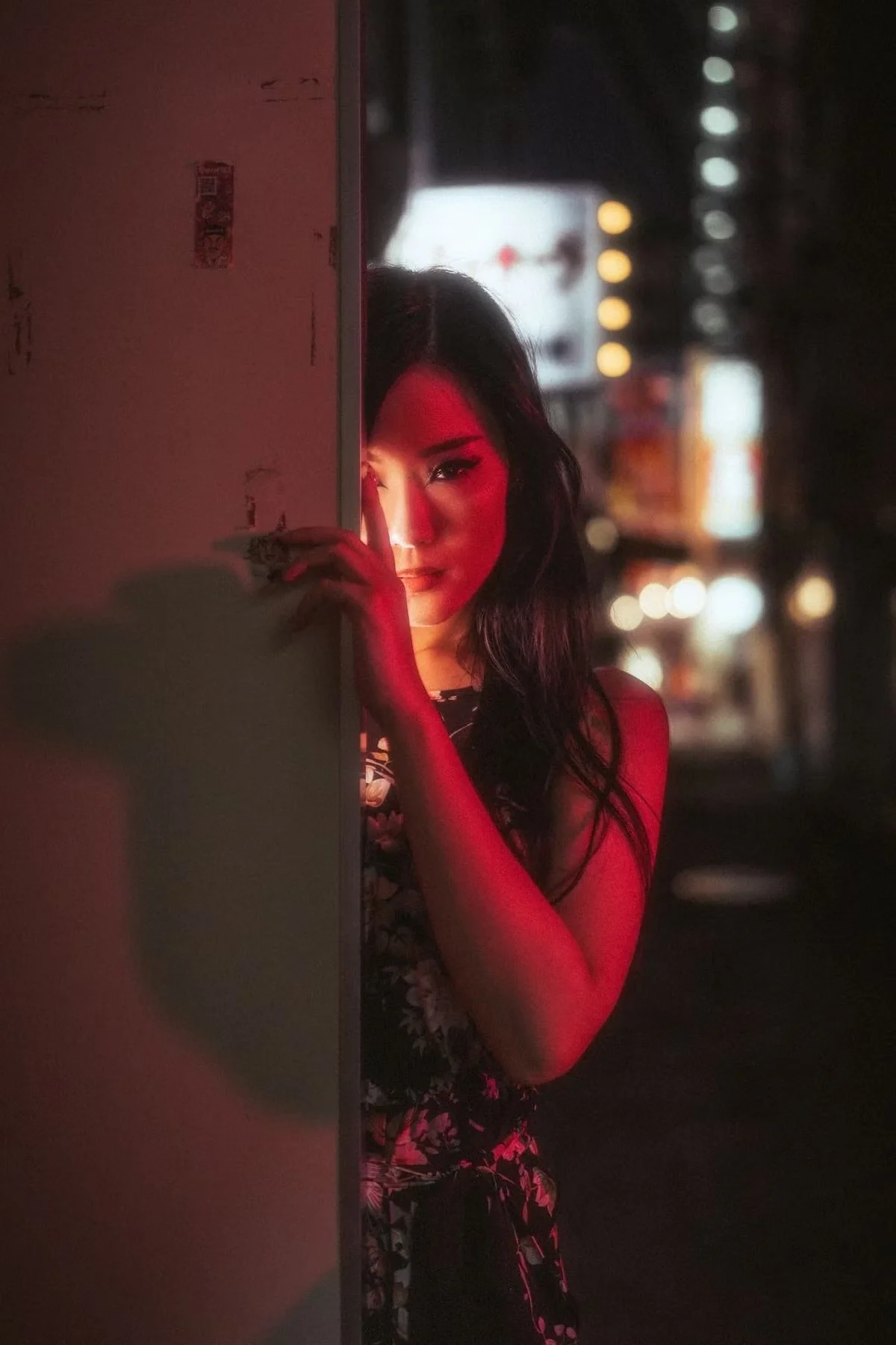 A Japanese woman peeking from behind a wall, illuminated by soft red and warm lights, with blurred city lights in the background. Shot by portrait photographer Deniz Demir.