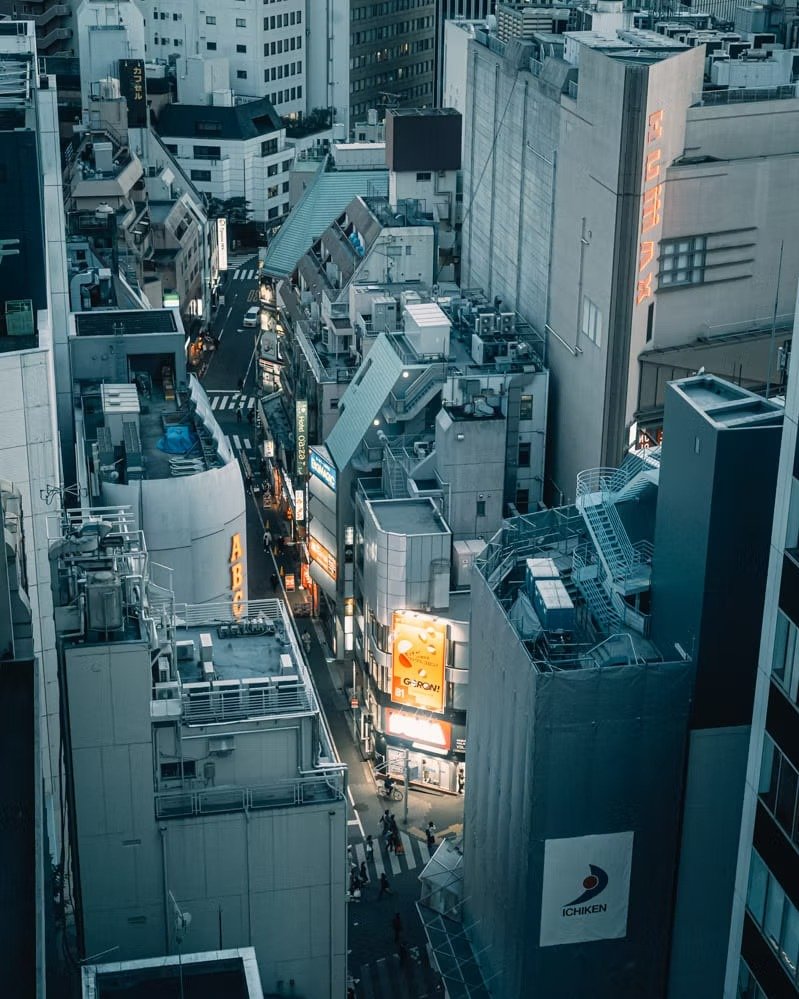 A view of a busy city street with tall buildings, illuminated signs, and pedestrians at dusk.