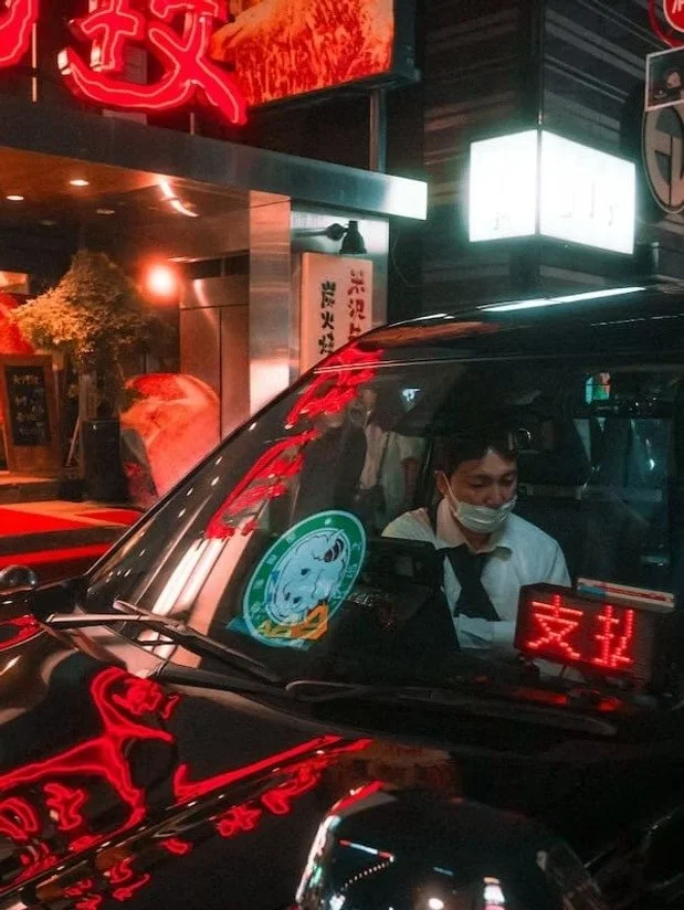 A black taxi cab with illuminated red and white signs, one of which is in Chinese characters. A person wearing a face mask is sitting in the driver's seat. The surroundings include city lights, signs, and reflections on the taxi's shiny surface.
