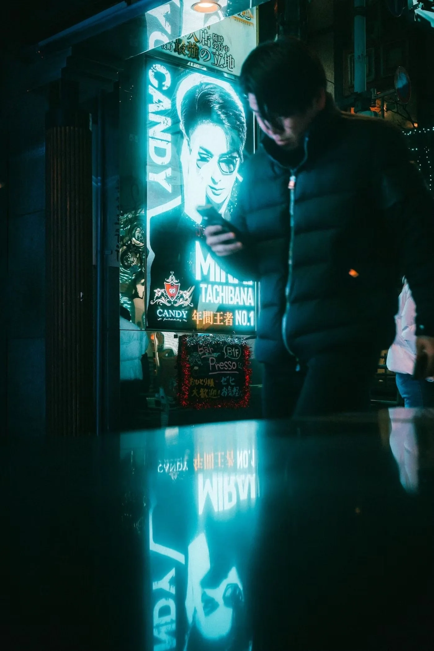 Night scene showing a neon sign with a woman's face and the words 'LADY TACIBANA', reflected on a shiny surface. A person in a dark jacket is walking past, partially blocking the view in Kabukicho Shinjuku.