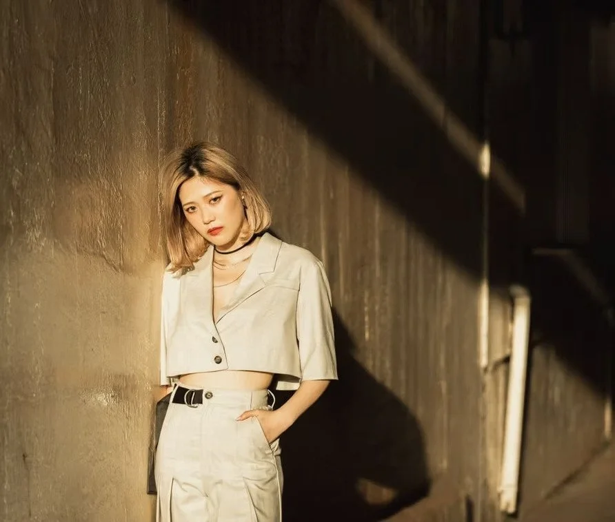 Young woman with shoulder-length blonde hair wearing a beige cropped blazer and matching pants, standing against a textured wall with shadowy lighting.