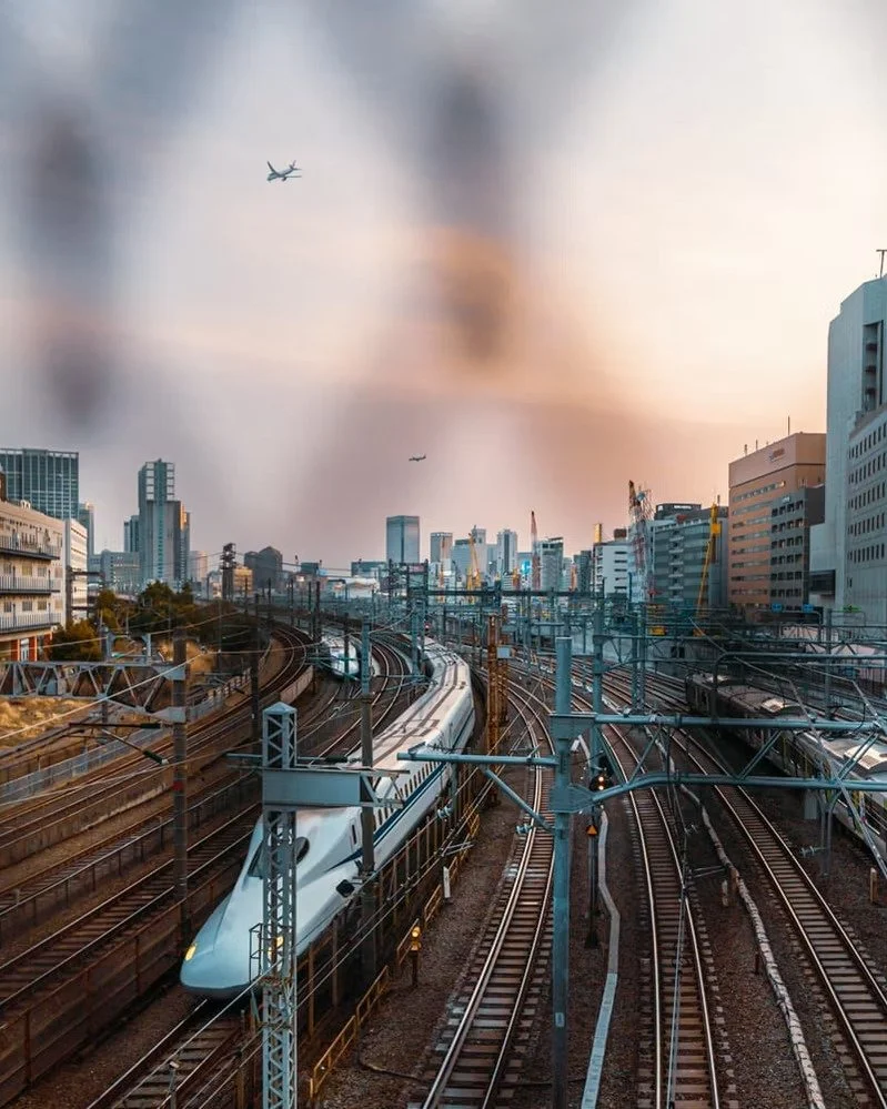 High-speed train on railway tracks in an urban city with skyscrapers and airplanes flying overhead during sunset.