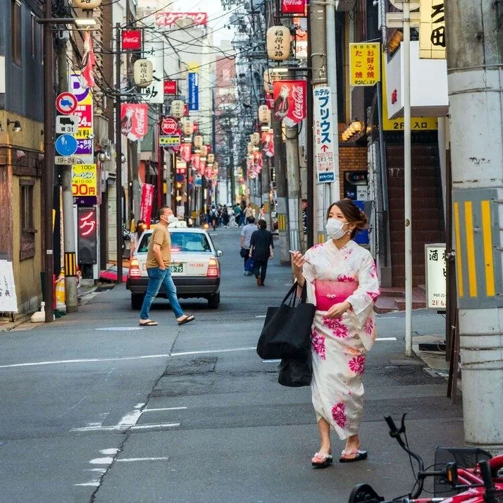 A woman in a pink and white kimono walks down a Tokyo street wearing a mask, carrying shopping bags. A fallen bicycle lies nearby, with cars in the background.