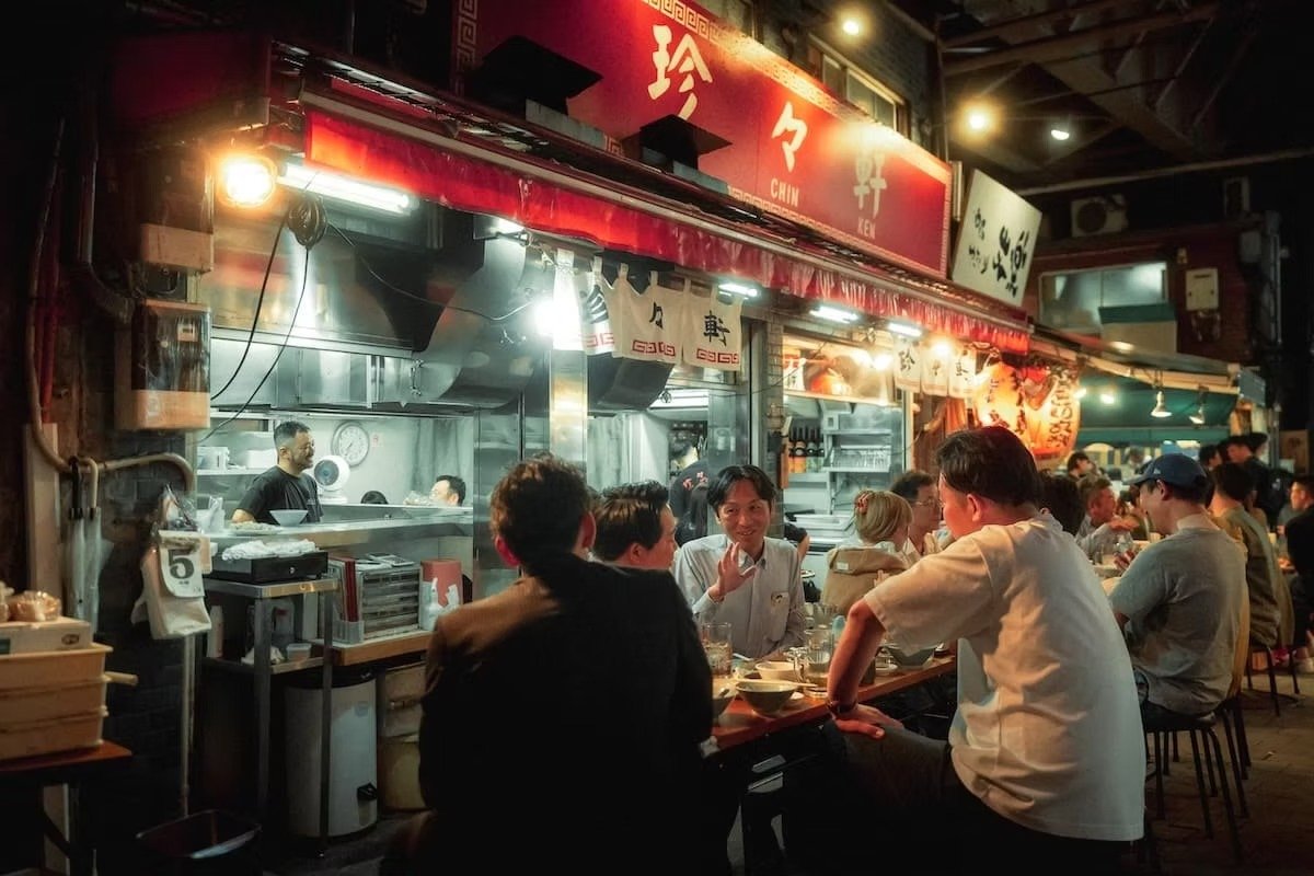 People dining at an outdoor Japanese restaurant at night with a busy open kitchen in the background. Ueno area by Tokyo street photographer Deniz Demir.