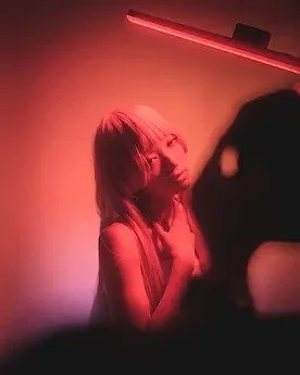A woman with shoulder-length hair and glasses posing with her head tilted in a dimly lit room with red and purple lighting, and a person taking a photo.
