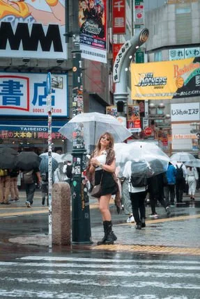 A woman stands under an umbrella in the busy Shibuya crossing area of Tokyo, surrounded by a crowd of people also carrying umbrellas. Bright neon signs and advertisements are visible in the background.